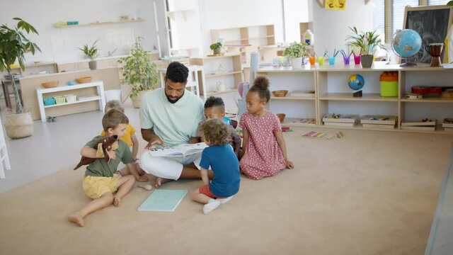 Group of small nursery school children with man teacher on floor indoors in classroom, reading book.