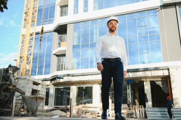 Successful engineer or architect, crane and building construction at backgrpound. Joyous businessman wearing helmet