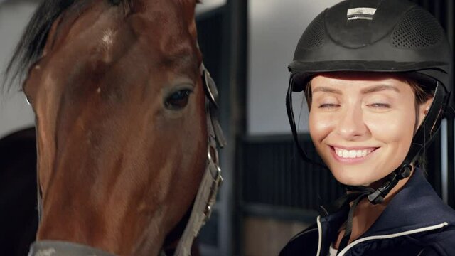 My Dear. Lovely Woman Worker Wearing Helmet Stroking To Her Brown Horse And Looking At The Camera While Spending Time At The Stable