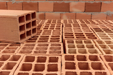 Pile of clay bricks used for building masonry house. In the image the bricks are laid out showing their holes, with one above them highlighted, and in the background an already built wall.

