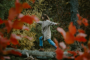 Woman in warm clothes doing impromptu yoga on a tree log in the forest framed by colourful tree...