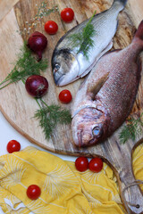 Uncooked Sea Bream fish on round wooden cutting board with cherry tomatoes, onion and spices. Food top view photo. Healthy eating concept. 