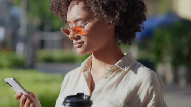 Black woman in stylish eyeglasses scrolling phone, drinking coffee during walk