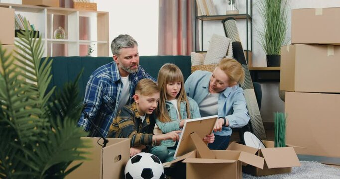 Adorable happy friendly smiling family with two kids sitting on the floor among carton boxes after relocation into new apartment and looking together on family portrait