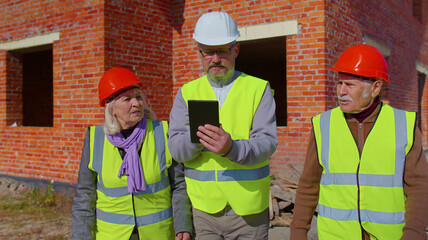 Diverse team of specialists taking near modern house. Real estate building project with senior civil engineer, architect, general worker discussing planning, development details on digital tablet