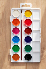 Children's watercolor paints and brushes in a palette on a wooden table. Hobby and leisure activity backgrounds