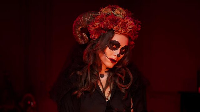 female halloween calavera image in the form of a skeleton with a sewn-up mouth, black eyes, horns, dry flowers on a beautiful Caucasian brunette woman in a dark room with red light