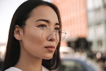 Close-up face of serious, confident asian woman in transparent round glasses looks into distance on blurred background. Long-haired brunette with natural makeup.