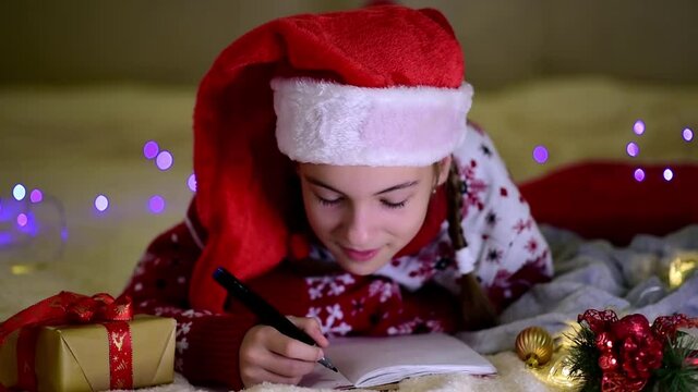 A teenage girl in Santa's red hat and red christmas jacket lies on the bed and writes a letter to Santa Claus. New Year. hildren's Christmas. Atmosphere. Home.