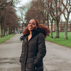 Beautiful African black woman walking in park in autumn. Beautiful young woman with brown skin. Smiling black woman with afro hair wearing black jacket.