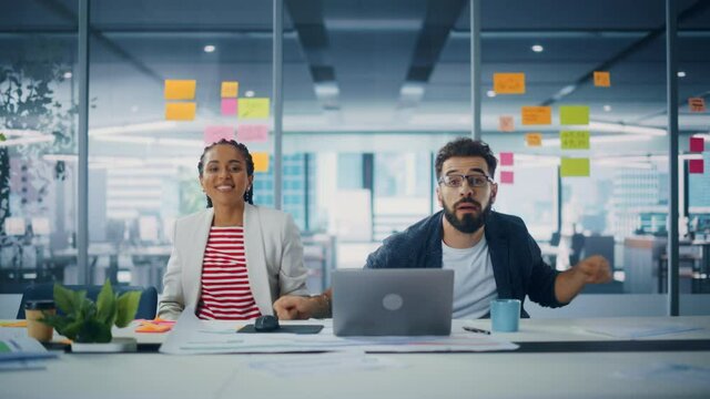 Two Happy Young Professionals in Office: Black Businesswoman and Hispanic Businessman Dance Together, Smile Looking at Camera, Sitting at Desk. Specialists Work in Creative Office, Celebrate Success
