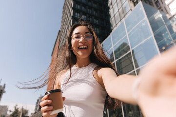 Close-up portrait brunette girl in white top looks into camera against background of panoramic windows of modern building. Makes photo holding glass of strong coffee in her hand.