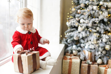 Fototapeta na wymiar Child girl dressed as Santa Claus near Christmas tree and gifts at home, winter New Year holiday