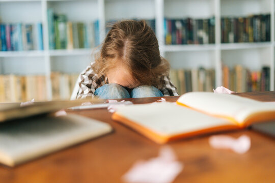 Front view of tearing unhappy primary child school girl sitting alone hugging knees in front of desk with difficult homework. Sad schoolgirl put head on knee being stressed with exam, selective focus
