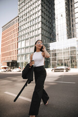  Full-length beautiful asian woman crosses road against backdrop of dark buildings. Makes important call on her way to work on spring day, taking cup of coffee with him.