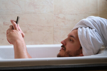 Young relaxed man using the phone lying in a bathtub filled with hot water.