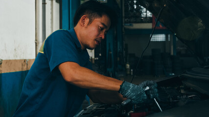 Fototapeta na wymiar Professional car mechanic screwing details of car engine on lifted automobile at repair service station. Skillful Asian guy in uniform fixing car at mechanics garage at night.