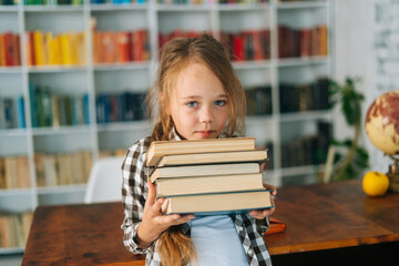 Medium shot portrait of adorable elementary child school girl holding stack of books in library at school looking at camera. Cute primary pupil schoolgirl standing on blurry background of bookshelves.