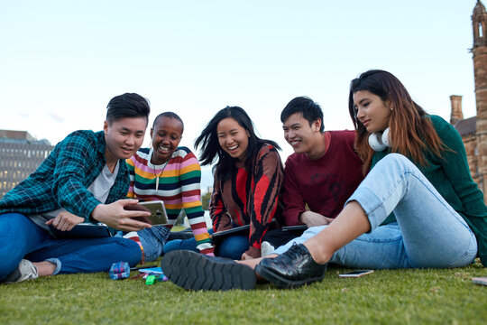 Group of young university students hanging out sitting on grass studying and using devices