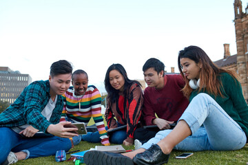 Group of young university students hanging out sitting on grass studying and using devices