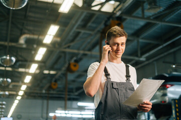 Low-angle view of smiling handsome young mechanic male wearing uniform holding clipboard and talking on mobile phone, standing in auto repair shop garage, with vehicle background, looking at camera
