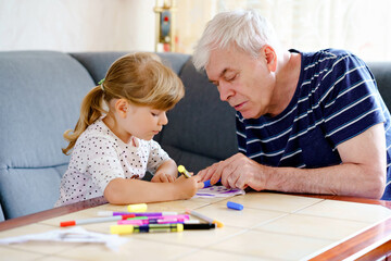 Cute little preschool girl and handsome senior grandfather painting with colorful felt pens and pencils at home. Grandchild and man having fun together, creative family. Indoor activity with child.