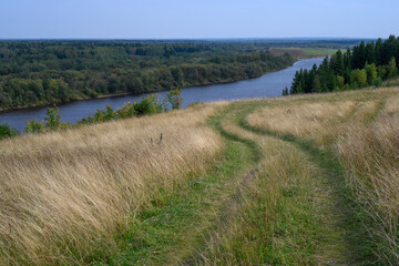Fototapeta premium Forest landscape. A winding dirt road on a mountain among tall yellow grass is directed into the distance to the Kolva River (Cherdyn, Russia). View from a height to the forest valley. Scenic road