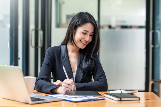 Beautiful young Asian businesswoman holding a pen using a tablet to work on accounting documents calculator laptop put on the table.