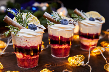 Christmas trifles in cups as a dessert, decorated with fresh blueberries and a vanilla stick, for a festive table on a background of glowing garlands