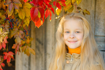 Naklejka na ściany i meble portrait of a smiling beautiful little blonde girl with long flowing hair in a not hat against a background of colorful vine leaves