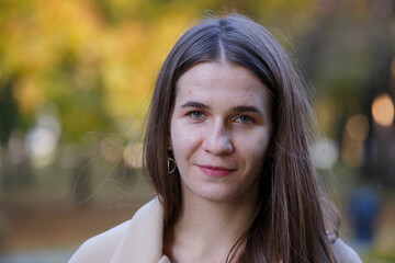 Portrait of a young pretty Caucasian girl with troubled skin wearing a beige coat against a blurry background of yellow leaves.