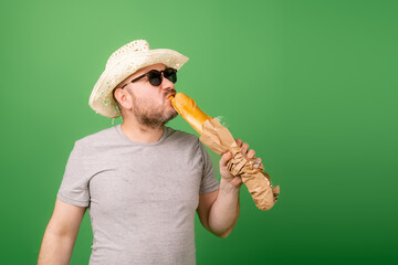 A man in a hat and sunglasses bites a French baguette on a green background.