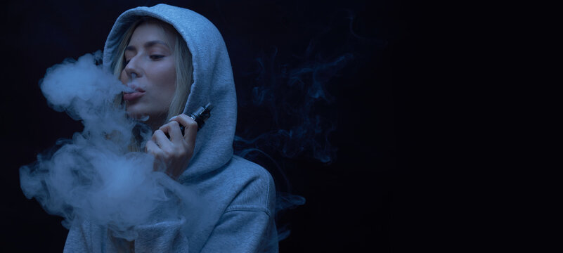 Portrait of beautiful blonde girl in gray hoodie smokes vape isolated on black studio background, cloud of steam smoke, mini hookah