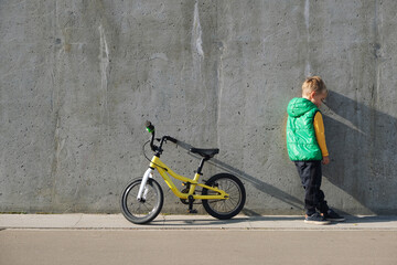 Fototapeta na wymiar Sad upset little boy standing near concrete wall with his bicycle on sunny day. Urban lifestyle.