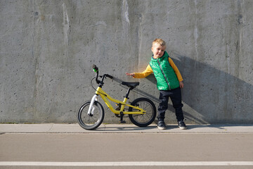 Fototapeta na wymiar Cheerful little child boy standing near concrete wall with his yelllow bicycle on sunny day. Urban lifestyle. Active kid in the city.