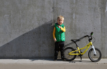 Fototapeta na wymiar Full length portrait of cheerful little child boy with yellow bicycle standing near concrete wall and pointing by finger on space for text. Urban lifestyle sportswear. Active kid in the city.
