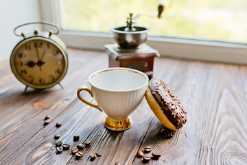 white kettle and cup on table. Breakfast by window. Chocolate donat next to scattered coffee beans.