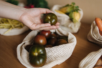 Female hand take tomato from canvas grocery bag. Vegetables in reusable eco cotton bags on wooden table. Zero waste shopping concept. Plastic free items