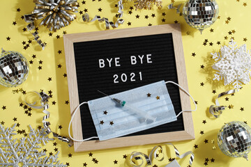 black letter board with text BYE BYE 2021 with decoration, syringe and mask on yellow background.