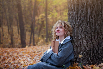 Beautiful girl reads a book in the autumn park. The girl has curly hair. Book in hand. Yellow autumn leaves. Copy space. Blurred background.