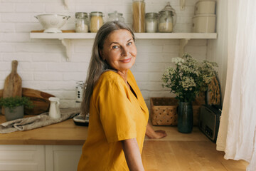 Lovely senior woman in yellow dress posing in countryhouse. Smiling positive female going to cook on kitchen, look from window. Weekends away, nature, interior, pure joy, growing old concept - Powered by Adobe
