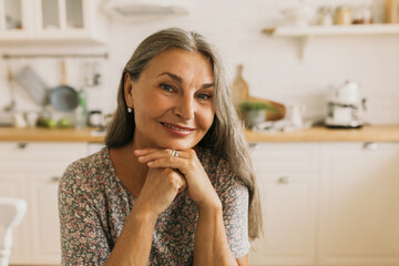 Beautiful Caucasian woman listening attentively to her interlocutor sitting in kitchen with chin on folded hands, looking at someone with interest, smiling, having conversation after dinner