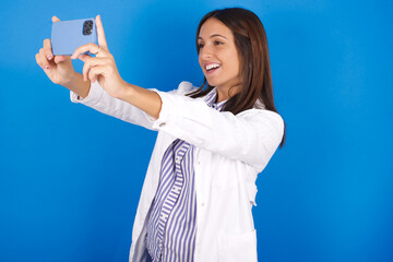 young european doctor girl wearing white medical gown ​on white background taking a selfie to...