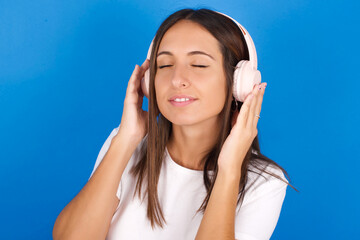 young european girl wearing white t-shirt on white background  with headphones on her head, listens to music, enjoying favourite song with closed eyes, holding hands on headset.