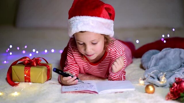 A little smiling girl in Santa's red hat and red christmas clothing lies on the bed and writes a letter to Santa Claus. New Year. Children's Christmas. Atmosphere. Home.