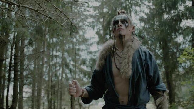 Worried young elf man with wooden stick walks along forest