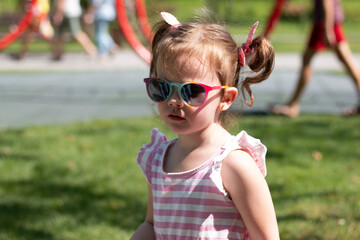 Active little beautiful girl with red pigtails, sunglasses and a striped dress runs around the playground