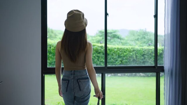4K slow motion of woman with luggage walking to watch the view through the window in a hotel room. Female enjoys hotel bedroom after check-in. Concept of travel and vacation.