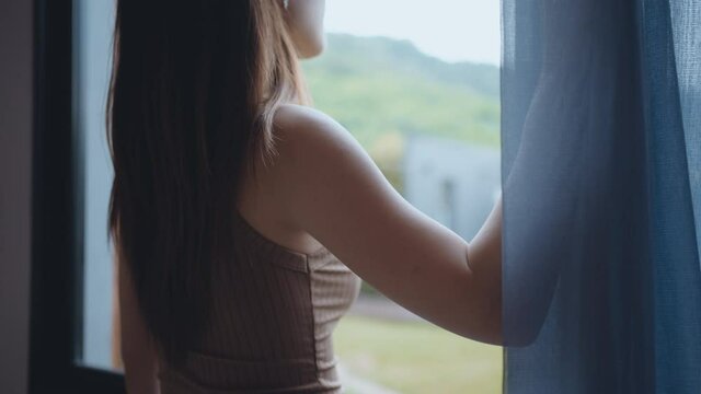 Asian woman opening curtains in hotel room looking out window after check-in for vacation trip