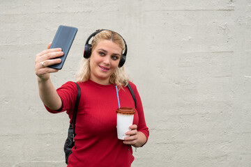 Portrait of an attractive young blonde haired woman with headphones taking a selfie with her cellphone and holding a plastic cup of coffee with a straw